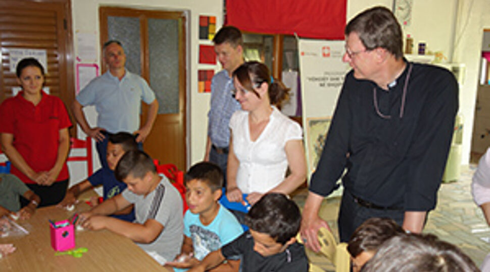 Kinder sitzen an einem Tisch und basteln. Kardinal Woelki und andere Teilnehmer der Delegation sind im Gespräch und schauen interessiert zu.